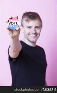 Portrait of a young man holding a VOTE badge up to the camera against pink background