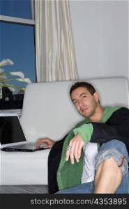 Portrait of a young man leaning against a couch and working on a laptop