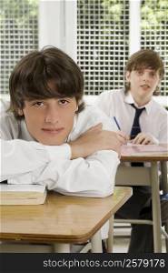 Portrait of a young man leaning on a desk with a teenage boy sitting behind him in a classroom