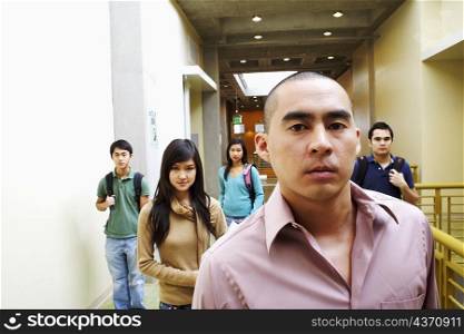 Portrait of a young man looking serious with two young women and two young men walking in the corridor behind him