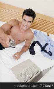Portrait of a young man lying on the bed using a remote control with a laptop in front of him