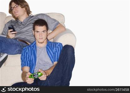Portrait of a young man playing video game with a remote control and another young man using a mobile phone behind him