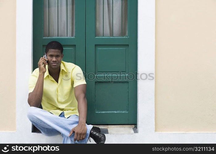 Portrait of a young man sitting in front of a door and talking on a mobile phone