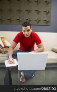 Portrait of a young man sitting in front of a laptop and writing on a spiral notebook