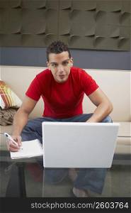 Portrait of a young man sitting in front of a laptop and writing on a spiral notebook
