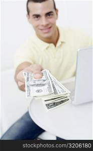 Portrait of a young man sitting in front of a laptop and holding one hundred dollar bills