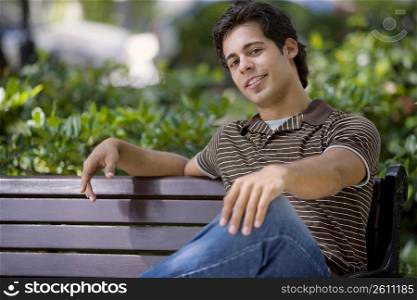 Portrait of a young man sitting on a bench with his hand on his knee and smiling