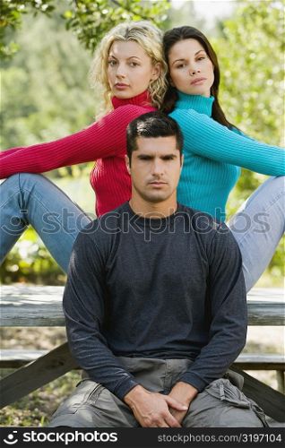 Portrait of a young man sitting on a bench with two young women sitting back to back behind him