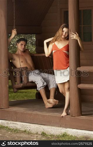 Portrait of a young man sitting on a porch swing with a young woman leaning against a column