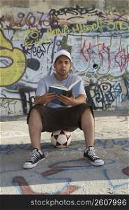 Portrait of a young man sitting on a soccer ball and holding a book