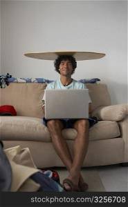 Portrait of a young man sitting with a surfboard on his head and using a laptop