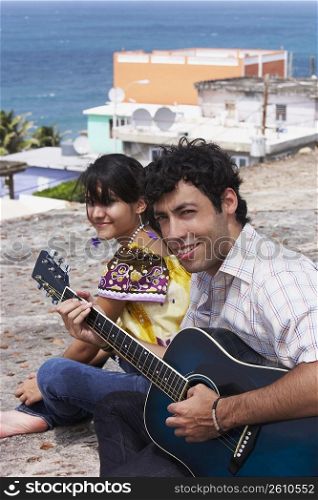 Portrait of a young man sitting with a young woman and playing a guitar