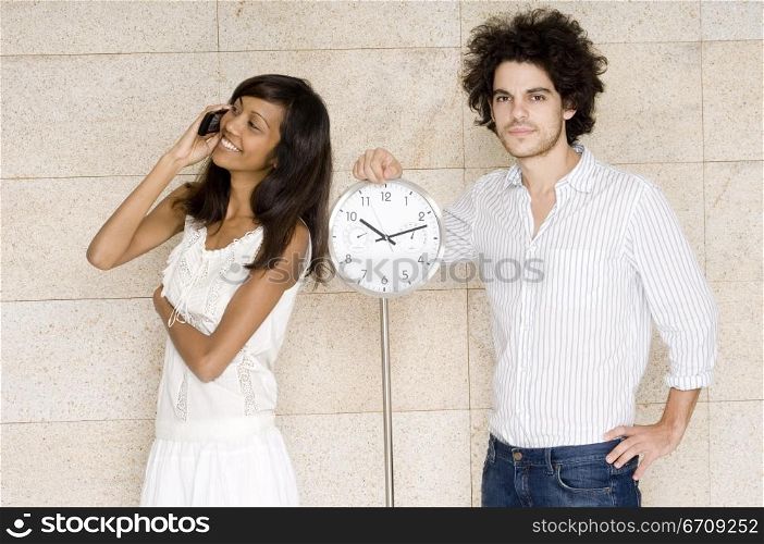 Portrait of a young man standing near a clock with a young woman talking on a mobile phone