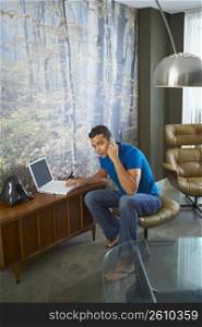 Portrait of a young man talking on a mobile phone while using a laptop