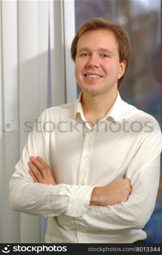 Portrait of a young man with kind smile and folded arms standing by the window with open blinds