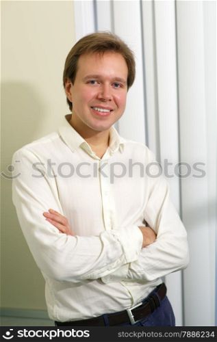 Portrait of a young smiling man posing with folded arms by the window with blinds