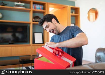 Portrait of a young surprised man opening a gift box while sitting on couch at home. Amazed and excited man.