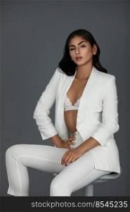 Portrait of a young white woman with wavy black hair and beautiful makeup posing by herself inside a studio with a grey background wearing a white suit with black high heels and white bra.