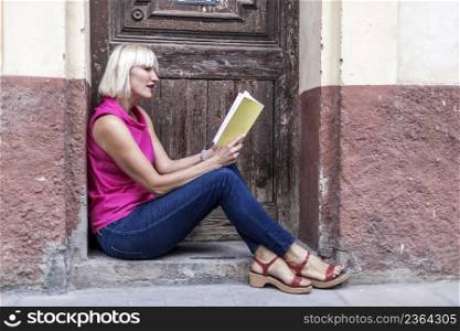 Portrait of a young woman concentrated reading book while sitting next to a house door during her recreation time, attractive Caucasian female enjoying rest and good day during autumn weekend
