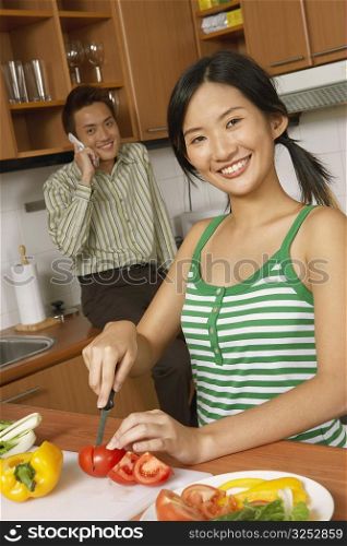Portrait of a young woman cutting vegetables in the kitchen with a young man talking on a mobile phone in the background