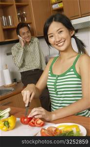 Portrait of a young woman cutting vegetables in the kitchen with a young man talking on a mobile phone in the background