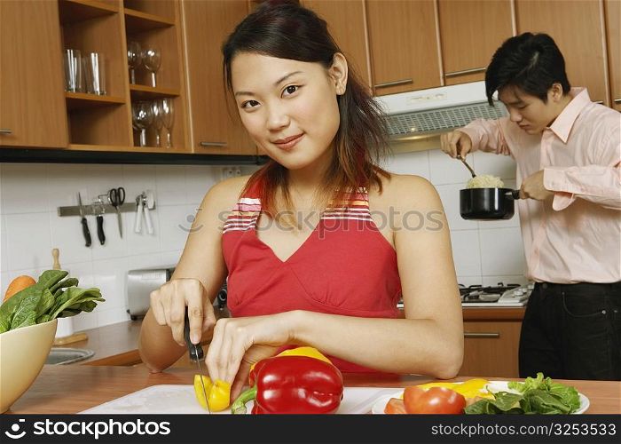 Portrait of a young woman cutting vegetables in the kitchen with a young man eating noodles in the background