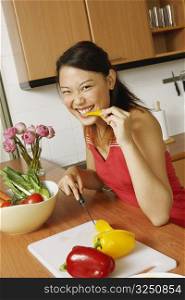 Portrait of a young woman eating a slice of yellow bell pepper at a kitchen counter