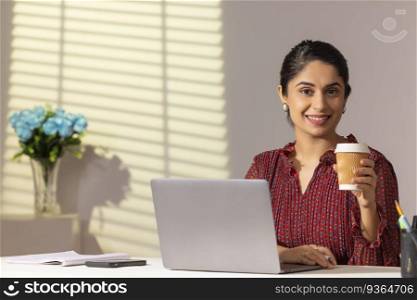 Portrait of a young woman executive looking at camera with holding cold coffee cup while sitting at her desk in office