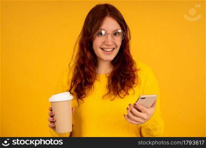 Portrait of a young woman holding a cup of coffee and using her mobile phone while standing against isolated background.
