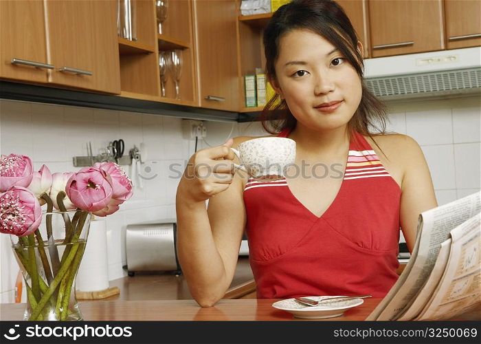 Portrait of a young woman holding a newspaper and a cup of tea at a kitchen counter