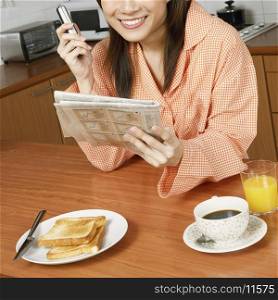 Portrait of a young woman holding a newspaper and a mobile phone at a kitchen counter