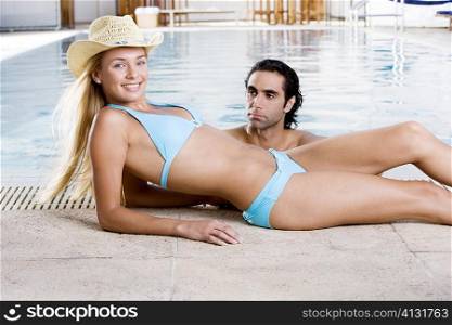 Portrait of a young woman lying down at the poolside with a mid adult man in the swimming pool