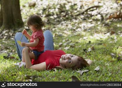 Portrait of a young woman lying on grass with her daughter sitting on her stomach