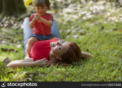 Portrait of a young woman lying on grass with her daughter sitting on her stomach