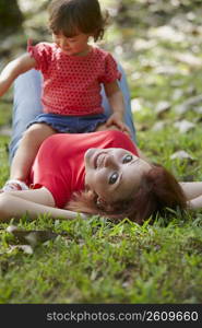 Portrait of a young woman lying on grass with her daughter sitting on her stomach