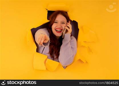 Portrait of a young woman pointing to the camera while talking on the phone through a hole in paper wall.