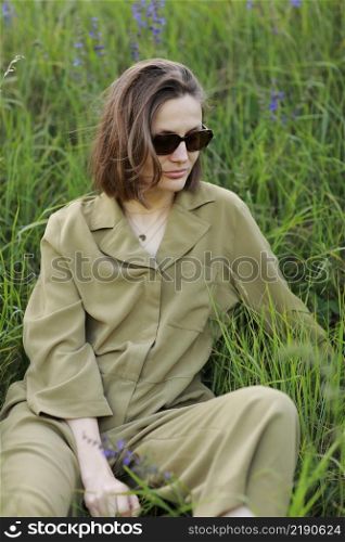 Portrait of a young woman sitting in a field on the spring grass among purple flowers. stylish girl wearing sunglasses enjoys Sunny spring weather. Natural beauty of a woman, natural cosmetics.. Portrait of a young woman sitting in a field on the spring grass among purple flowers. stylish girl wearing sunglasses enjoys Sunny spring weather. Natural beauty of a woman, natural cosmetics