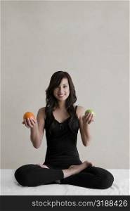 Portrait of a young woman sitting in a yoga position and holding an orange and an avocado in her both hands