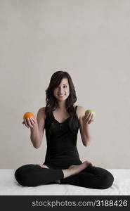 Portrait of a young woman sitting in a yoga position and holding an orange and an avocado in her both hands