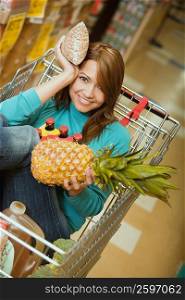 Portrait of a young woman sitting in shopping cart and holding a pineapple and a packet