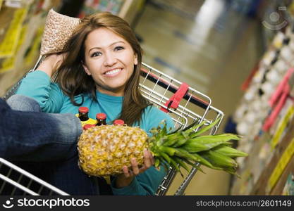 Portrait of a young woman sitting in shopping cart and holding a pineapple