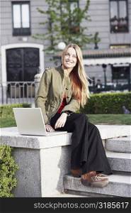 Portrait of a young woman sitting on a ledge and using a laptop