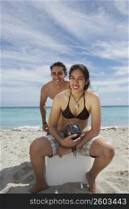 Portrait of a young woman sitting on an ice box on the beach with a young man standing behind her