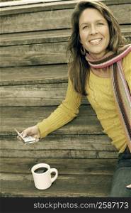 Portrait of a young woman sitting on the wooden steps with a coffee cup and holding a personal data assistant