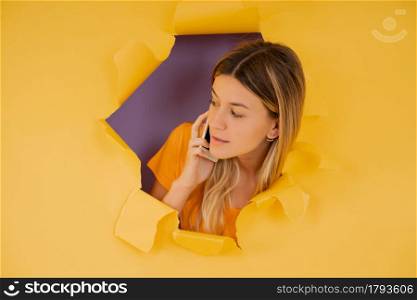 Portrait of a young woman talking on the phone through a hole in paper wall.