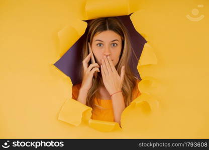 Portrait of a young woman talking on the phone with shocked expression on her face while standing through a torn hole in paper wall.