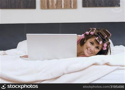 Portrait of a young woman wearing hair rollers and hiding herself behind a laptop on the bed