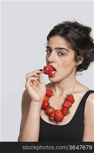 Portrait of a young woman wearing strawberry necklace as she eats one piece over gray background