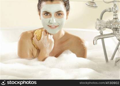 Portrait of a young woman with a facial mask holding a bath sponge in a bathtub