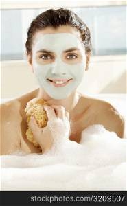 Portrait of a young woman with a facial mask holding a bath sponge and smiling